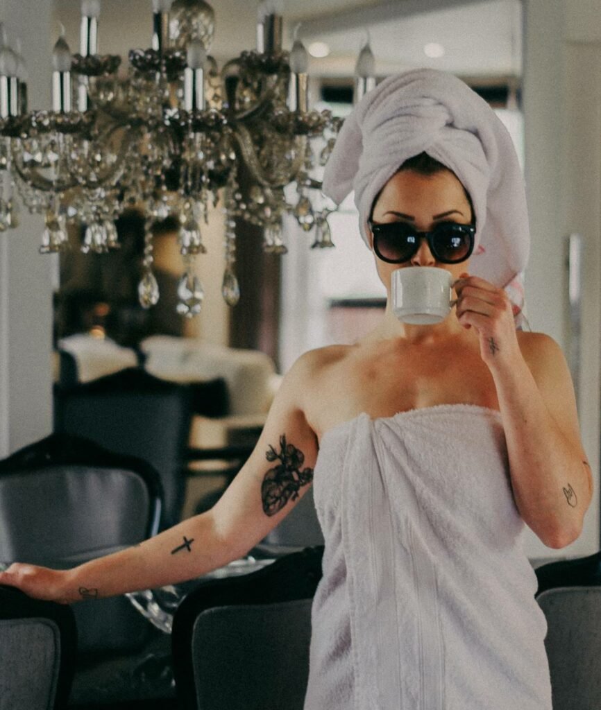 Mulher branca, com tatuagens, de toalha em sua sala bebendo uma xícara de chá com uma toalha no cabelo também.