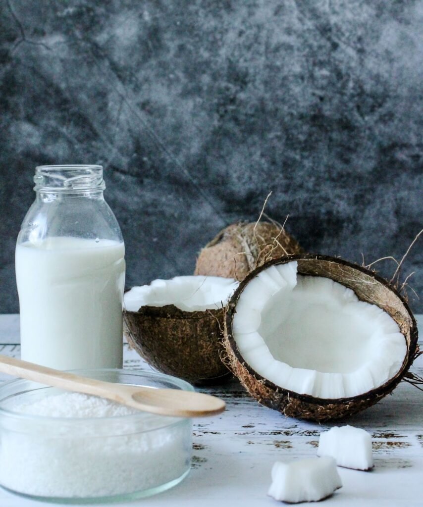 a imagem mostra um coco seco (marrom) aberto, seu interior é branco. Há também um pote de vidro redondo e baixo com coco ralado dentro, com uma colher de pau marrom claro em cima. À esquerda do coco e atrás do pote de vidro, há uma garrafa de vidro aberta com leite de coco dentro. Todos esses ingredientes estão em cima de uma mesa de madeira pintada de branca, ao fundo há uma parede de mármore.