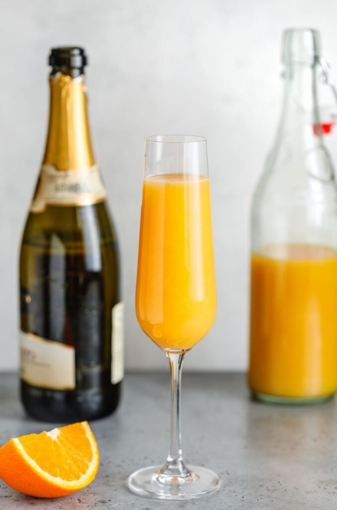 Taça com mimosa dentro, uma bebida amarela). Há uma fatia de laranja, um espumante e uma garrafa com mimosa ao fundo.