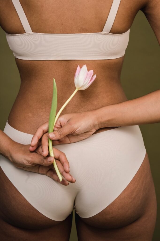 Mulher negra, de costas, segurando uma flor rosa claro. Ela usa uma calcinha e sutiã na cor branca.