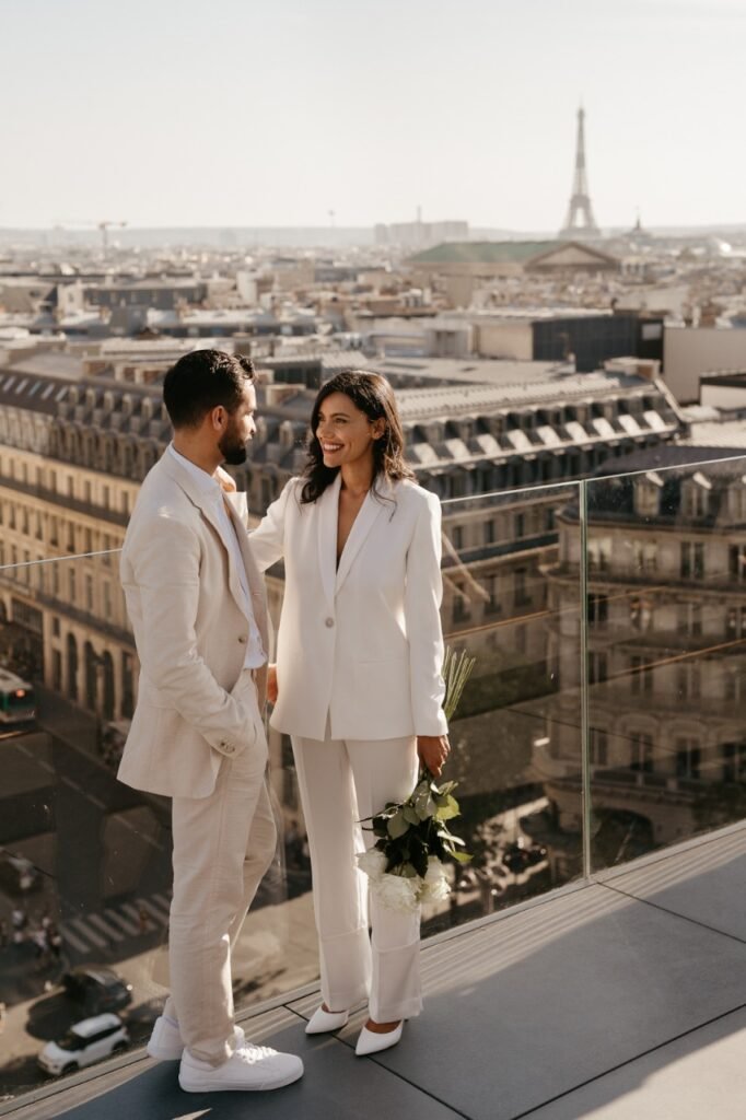 Casal na sacada de um hotel, é possível ver o cenário da cidade de Paris, pois a Torre Eiffel está ao fundo.  