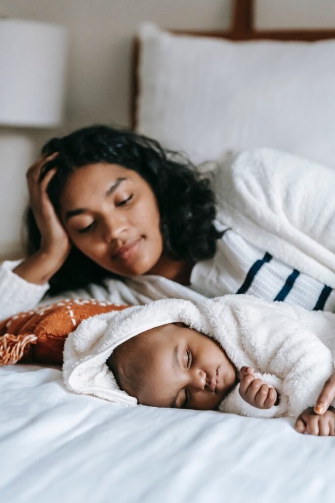 Mulher negra com seu bebê, ambos deitados na cama. Ela está o observando, enquanto ele dorme.