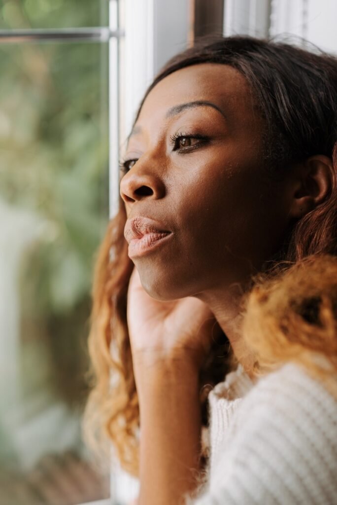 Mulher negra, de olhos castanhos e cabelo castanho com ombre hair loiro, observando a janela. 