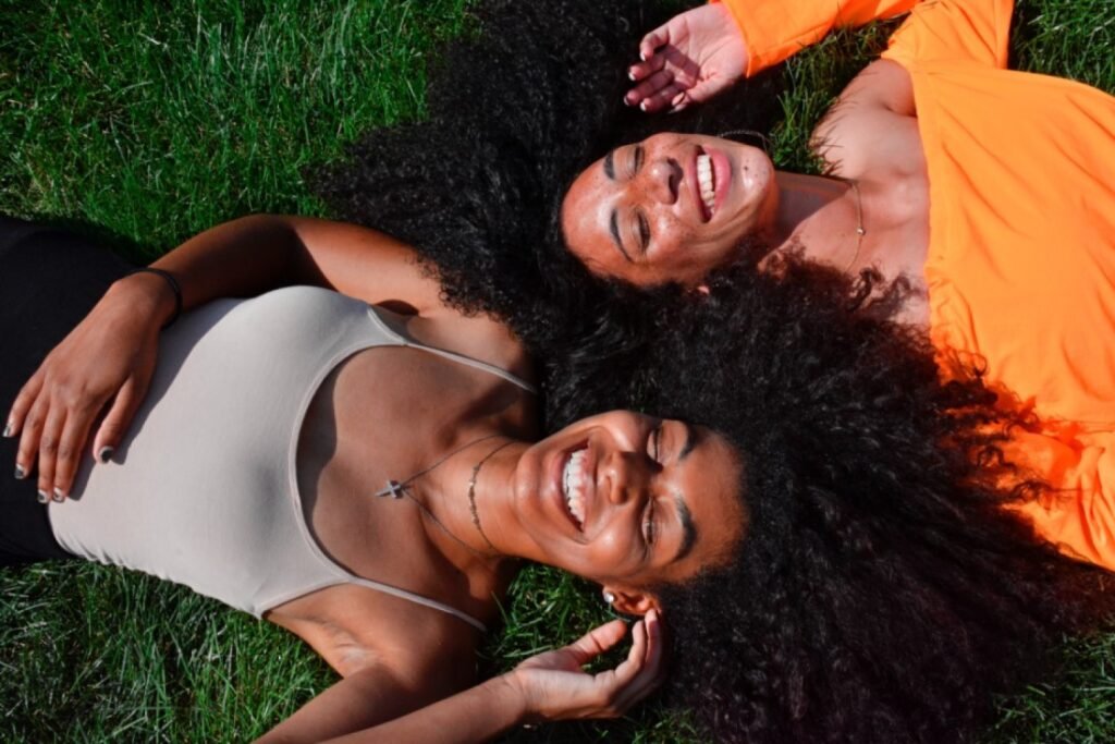 Duas mulheres negras, deitadas na grama sorrindo. As duas possuem cabelos cacheados castanhos e estão com a luz do sol em seus rostos. 