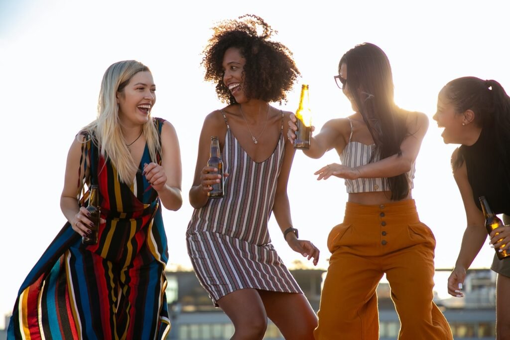 Quatro mulheres estão sorrindo e cada uma delas segura uma garrafa de cerveja. Elas parecem estar no terraço de um prédio. 