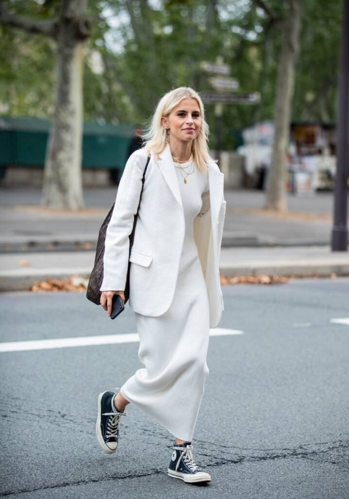 Uma mulher branca, de cabelo loiro liso na altura dos ombros, está em uma rua. Ela veste um vestido longo branco, blazer branco e tênis all star preto.