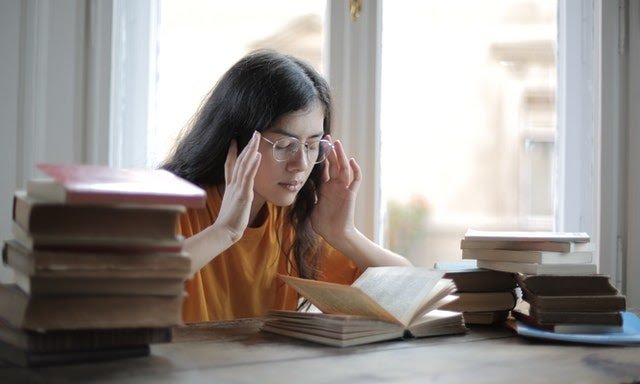 Mulher branca de cabelos castanhos longos. Ela está sentada de frente para uma mesa, com diversos livros. Está segurando a lateral dos óculos de grau, com dor de cabeça.