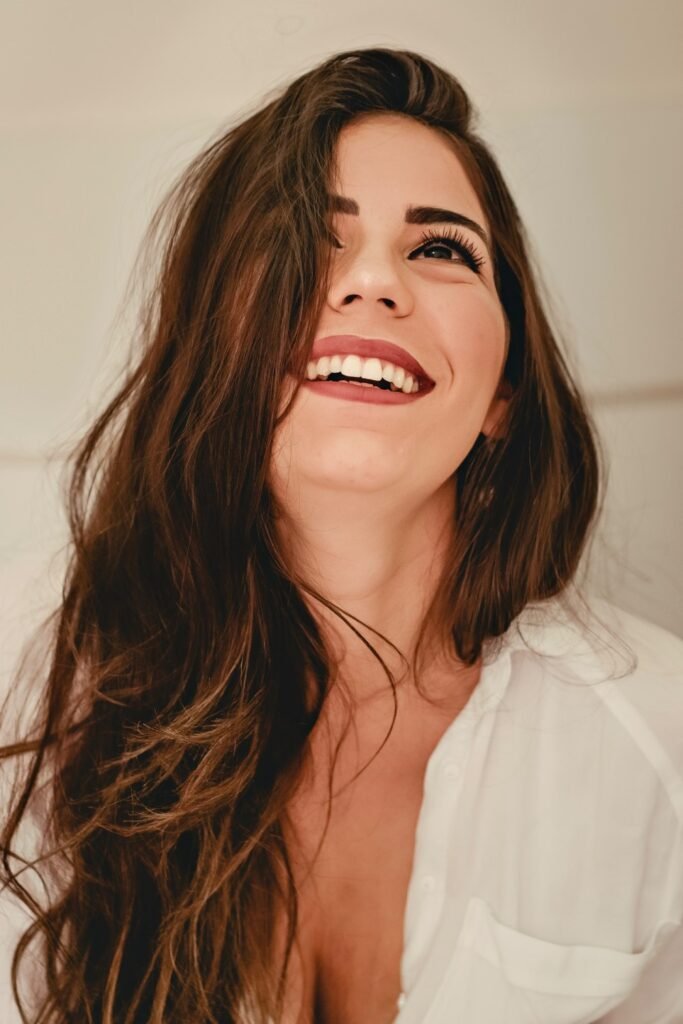 Uma mulher branca, de cabelos castanhos-escuros lisos, está olhando para cima e sorrindo. Ela veste uma camisa branca.