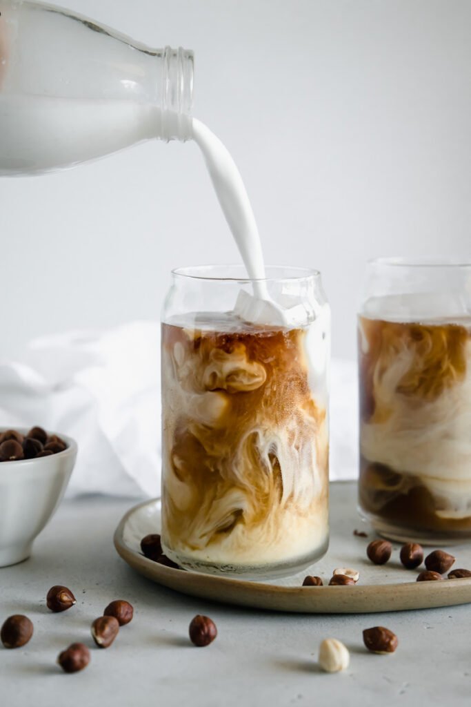 Uma garrafa de vidro com leite despeja um pouco da bebida em um copo de vidro, que contém um pouco de café. Ao redor é possível ver mais um copo de vidro com a bebida e alguns grãos de café sobre a mesa. 