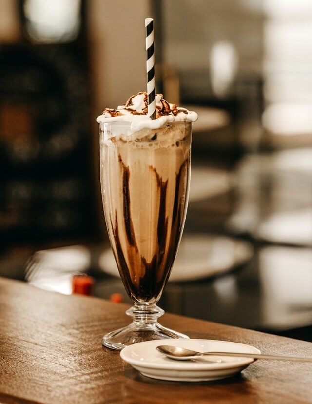 Uma taça de milk shake aparece sobre uma mesa de madeira. Também há uma colher de sobremesa de metal sobre um pires branco.  