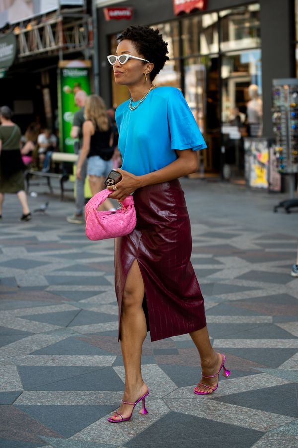 Mulher negra, de cabelo cacheado preto curto. Ela está em um cenário urbano, na calçada, com uma t-shirt azul-claro e uma saia midi vermelha com fenda. 