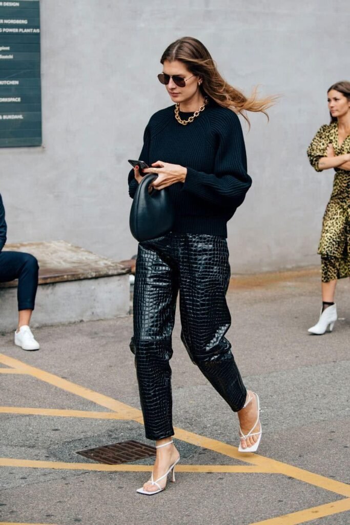 Uma mulher branca de cabelo castanho-claro liso está em uma calçada. Ela veste blusa de manga longa, calça de couro preta e sandália de salto branca. 