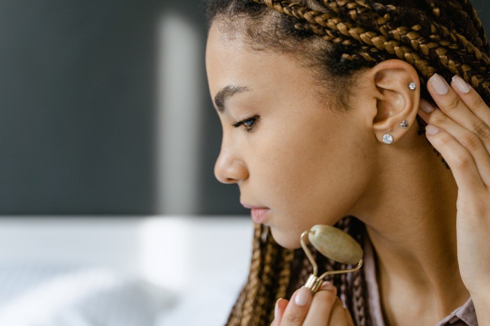 Uma mulher negra, com tranças loiras nos cabelos está de perfil. Ela usa um massageador facial de pedra de jade.