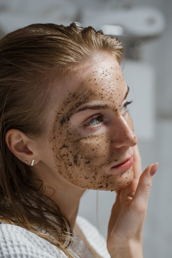 Uma mulher branca, de cabelo loiro liso molhado, está fazendo uma exfoliação facial com um produto preto para evitar os poros dilatados. 