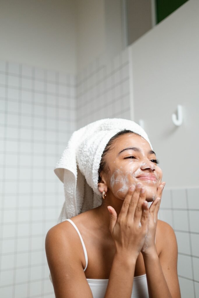 Uma mulher negra está com os cabelos enrolados em uma toalha. Ela espalha um creme branco em seu rosto. 