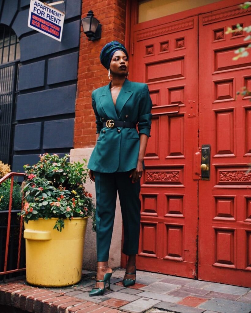 Mulher negra usando turbante, conjunto de alfaiataria e salto em tom de verde escuro com um cinto preto e símbolo de marca no meio. Ao fundo uma porta vermelha e ao lado um vaso amarelo com plantas com flores.