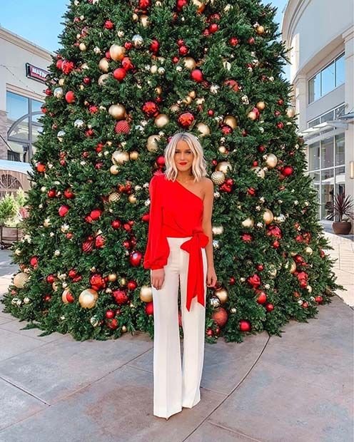 Mulher branca e loira em frente a uma árvore de Natal usando uma blusa de uma manga comprida com um laço na cintura e uma calça branca.