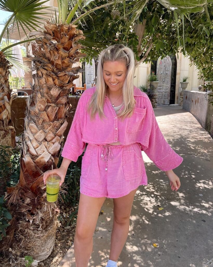 Uma mulher branca, loira de cabelos lisos, usa um look de verão: blusinha e shorts rosa. Eça está em frente a um coqueiro.