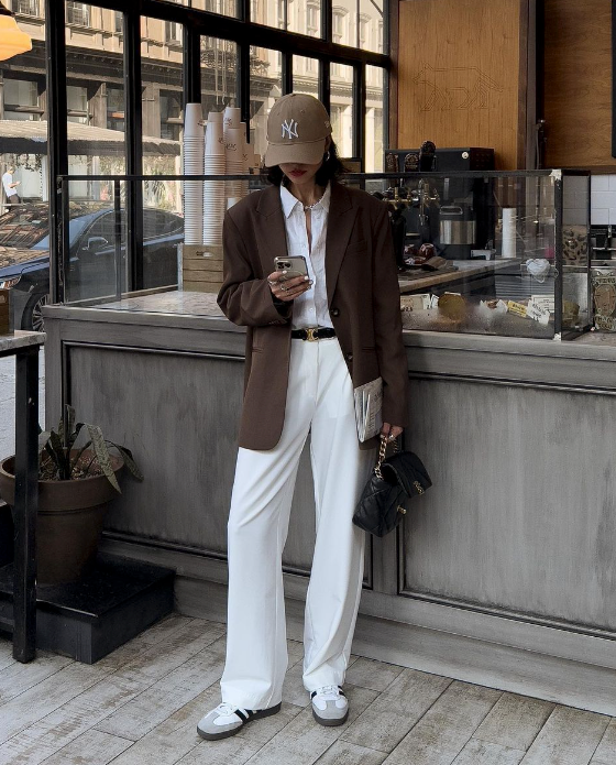 Jovem coreana vestindo calça social off-white, camisa branca, blazer marrom, tênis branco e boné marrom