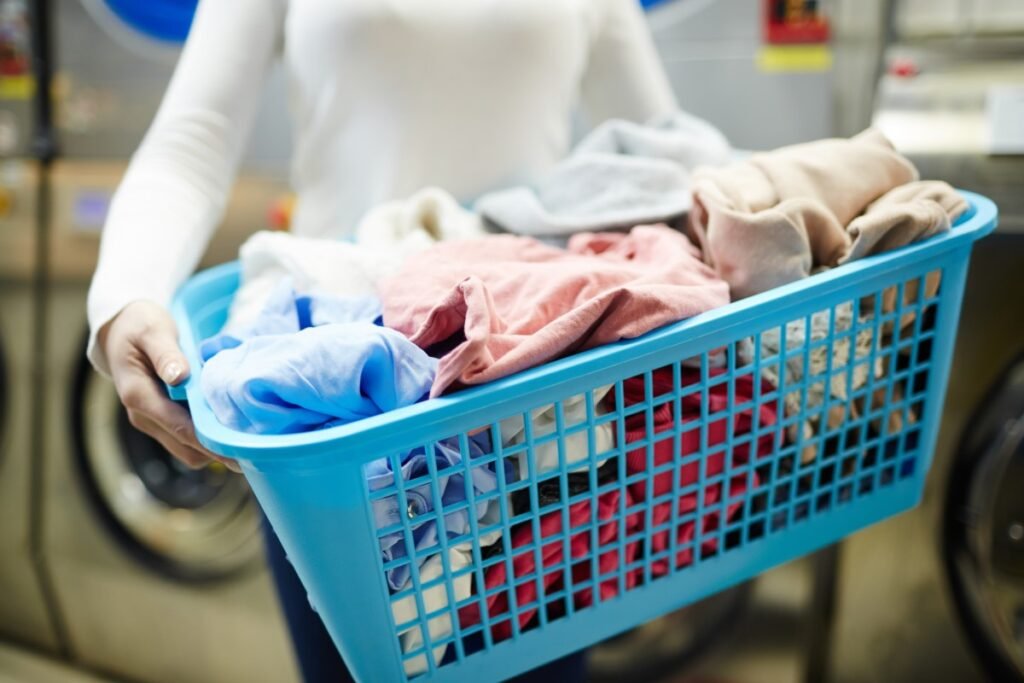 Em uma lavanderia, uma mulher de blusa longa branca e jeans segura o cesto de roupas azul com algumas roupas dentro.