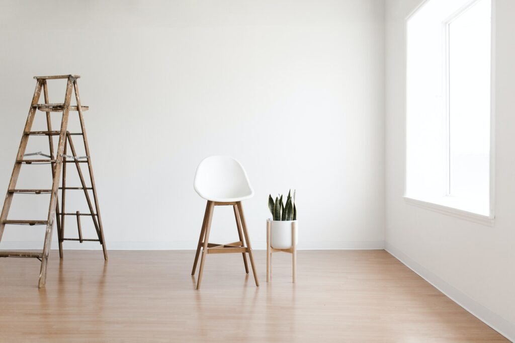 Alt text: Uma sala minimalista com paredes e janela brancas, com um banco com pernas de madeira e assento branco, um vaso branco em um suporte de madeira e uma escada aberta, também de madeira.