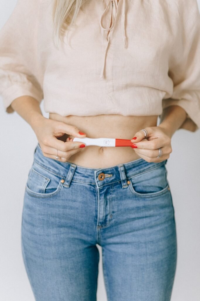 Uma mulher loira, usando jeans, uma blusa dobrada e esmalte vermelho, segura um teste de gravidez branco e vermelho próximo a barriga em um fundo branco. Os diferentes tipos de DIU ajudam na prevenção da gravidez. 