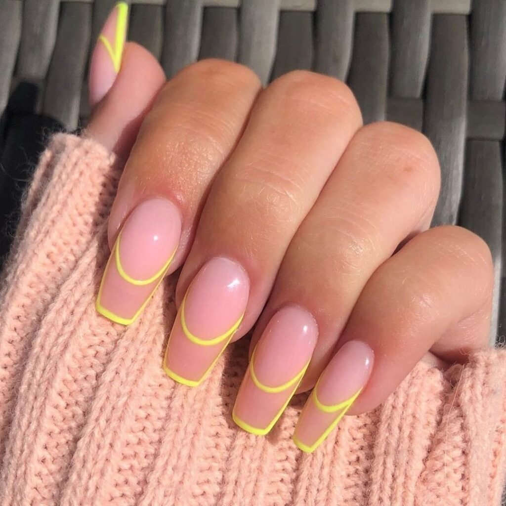 Mão branca com unhas quadradas pintadas de rosa-claro e detalhes geométricos em amarelo.