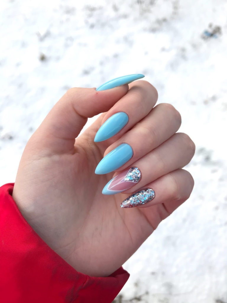 Mãos brancas com unhas stiletto pintadas de azul e o anelar misturado brilhos, nude e azul, e a unha mínima pintada com os mesmos brilhos.