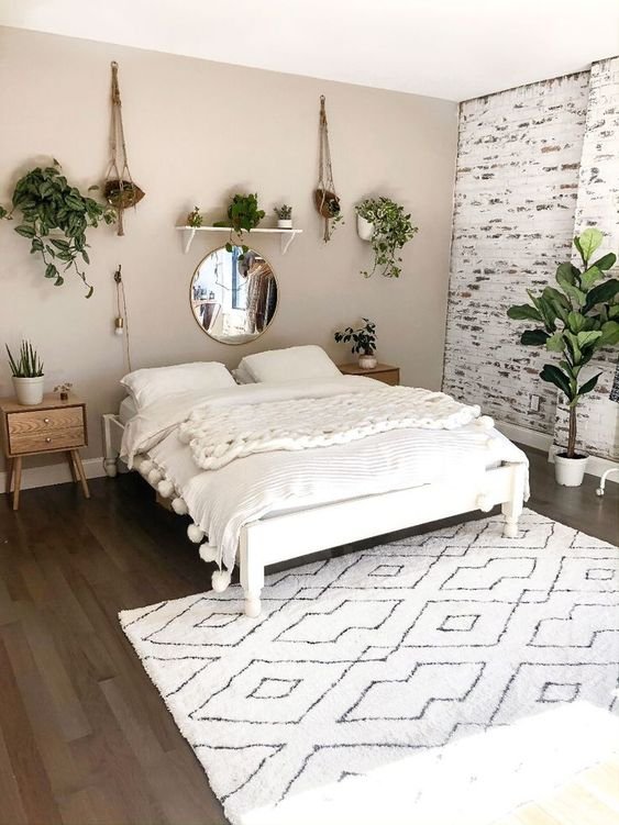 Foto de um quarto boho. O chão é de madeira escura, há um tapete étnico em branco e preto, uma cama branca com roupa de cama branca e mantas por cima, as paredes são em tons claros, com plantas e um espelho redondo.