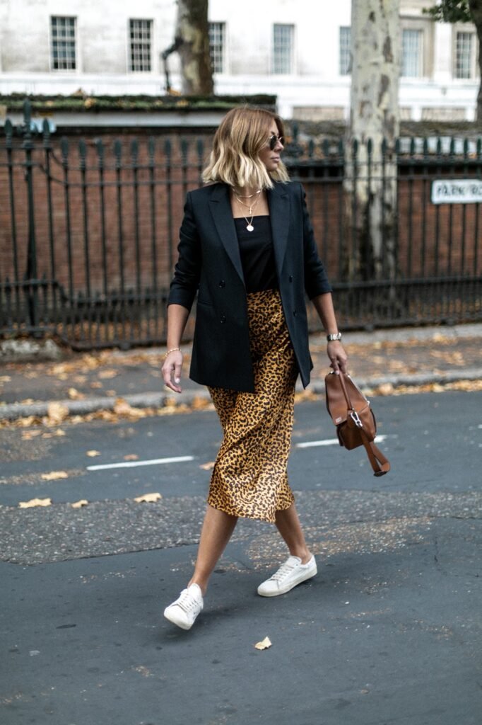 Uma mulher branca, loira, está andando em uma rua. Ela veste um blazer preto, blusinha preta e saia midi com estampa de onça.