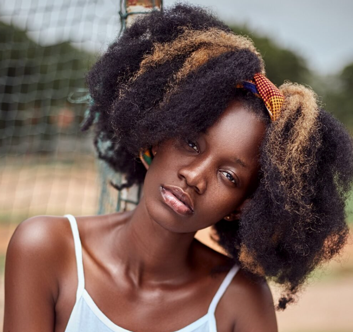 Mulher negra com cabelo crespo repartido no meio com mechas loiras e uma tiara 