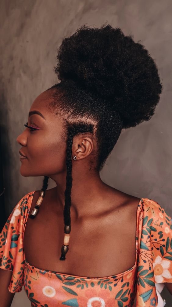 Mulher negra usando um dos tipos de penteados para cabelo crespo: duas tranças laterais e o restante do cabelo preso no topo da cabeça. Ela veste uma blusa com mangas largas de cor laranja e estampa floral.