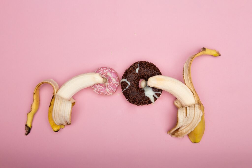 Alt text: Em um fundo rosa, estão dois donuts, um rosa pequeno e um marrom maior, com duas bananas descascadas sobre eles. A ginástica íntima é uma forma de trazer mais prazer para a mulher na relação sexual.