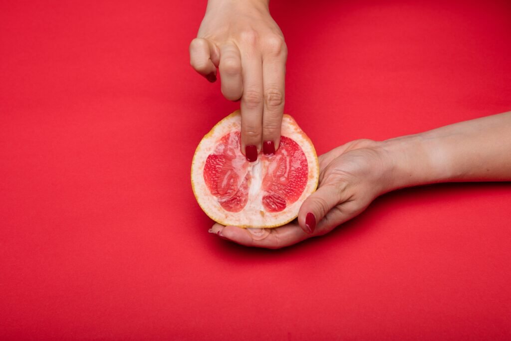 A imagem mostra as mãos de mulher com unhas pintadas de vermelho segurando uma toranja e pressionando-a com dois dedos, como pode ser feito na massagem yoni.