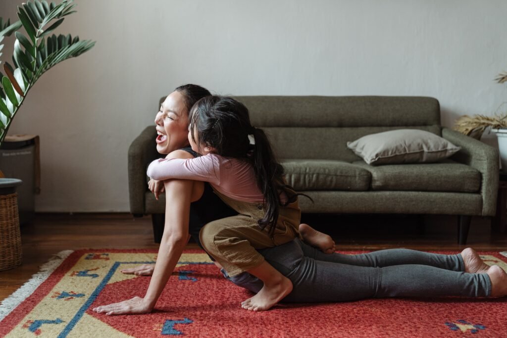 Mãe e filha abraçadas e rindo enquanto a mãe faz yoga no tapete da sala, colocando em prática uma das dicas de autocuidado.