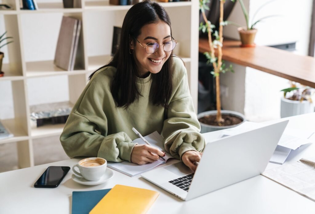 Mulher trabalhando no computador, sorrindo e fazendo anotações em um caderno